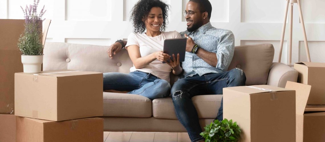Happy african couple husband and wife choose house removal service or search renovating ideas sit on sofa with boxes, smiling black renters owners tenants use digital tablet on moving day in new home
