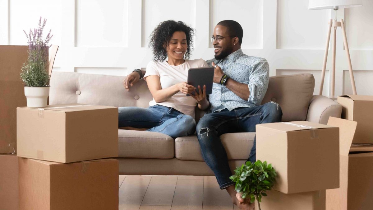 Happy african couple husband and wife choose house removal service or search renovating ideas sit on sofa with boxes, smiling black renters owners tenants use digital tablet on moving day in new home
